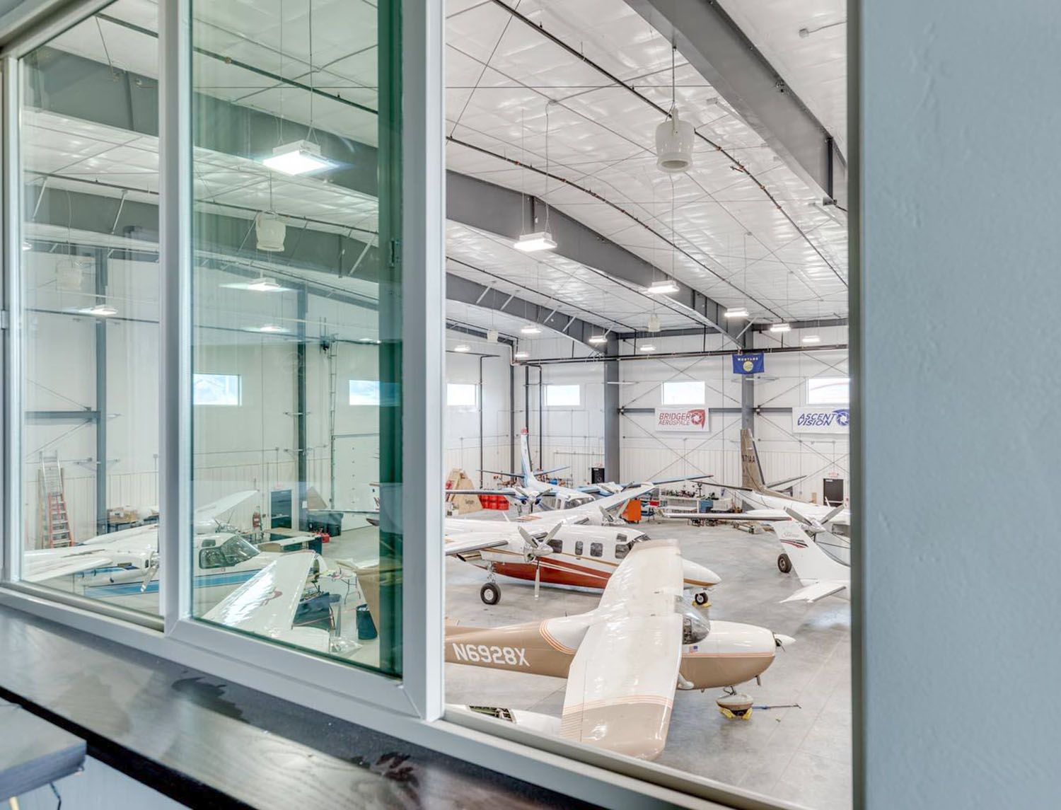 A view through a window into a brightly lit aircraft hangar containing several small, single-engine airplanes