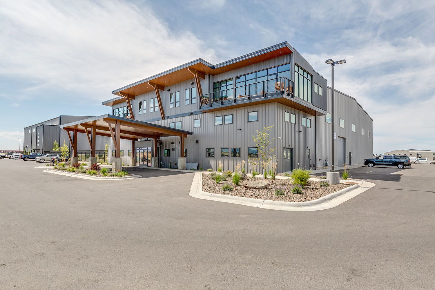 A modern, three-story grey office building with wooden accents, a covered entrance, and a parking lot under a cloudy sky