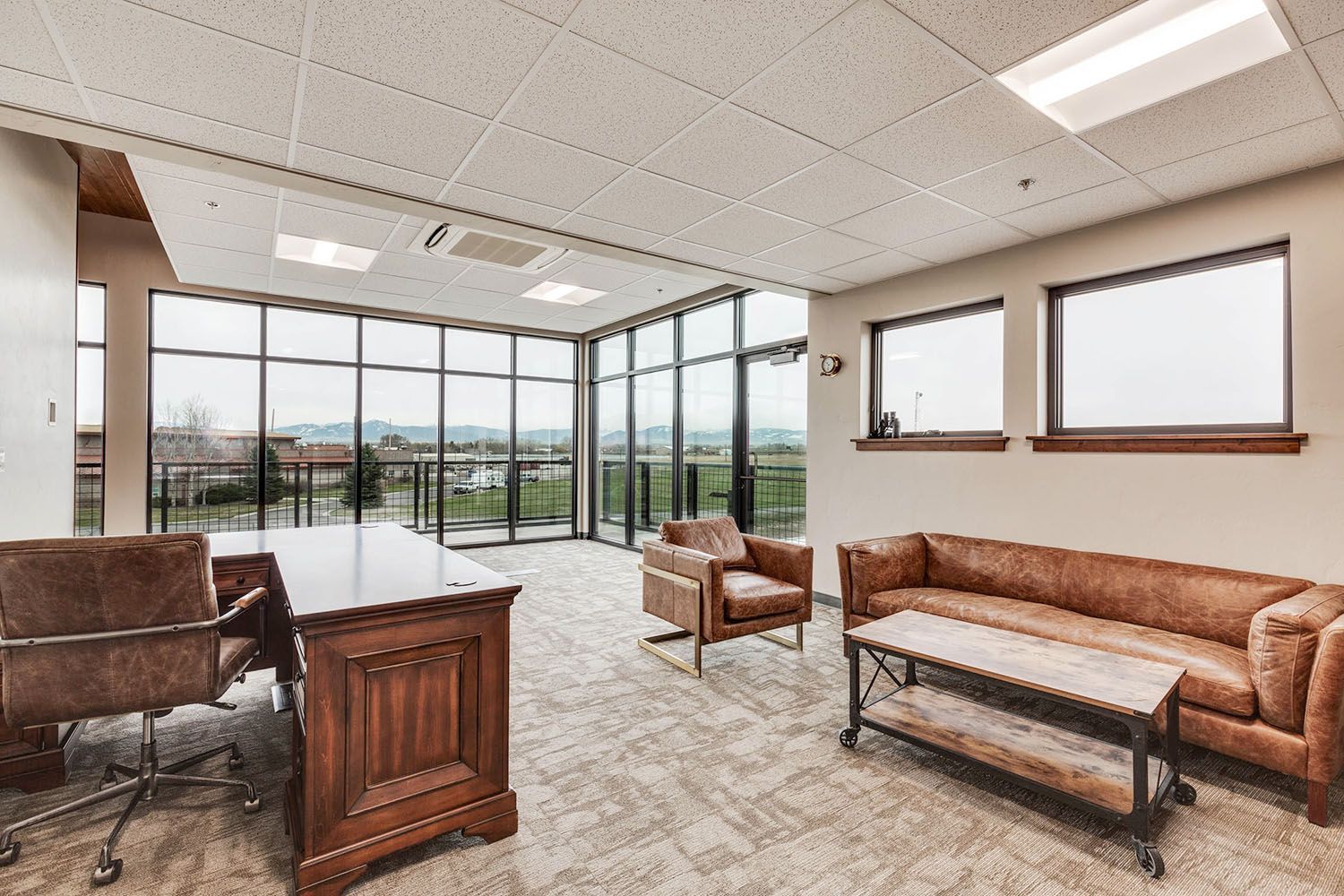 A modern professional office with a wooden desk, leather chair, sofa, and coffee table against large windows