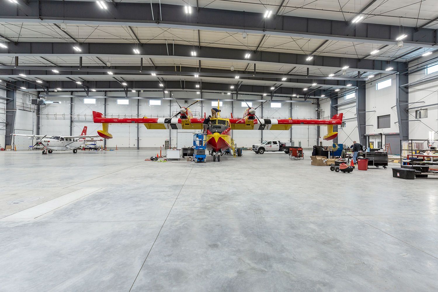 A bright yellow and red aerial firefighting plane sits inside a large, spacious hangar alongside another small aircraft