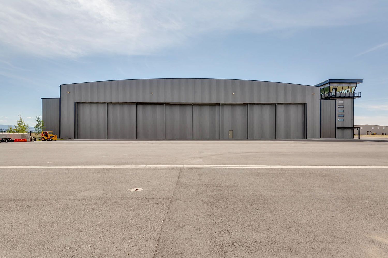 A large gray metal aircraft hangar with sliding doors and an attached control tower under a bright blue sky