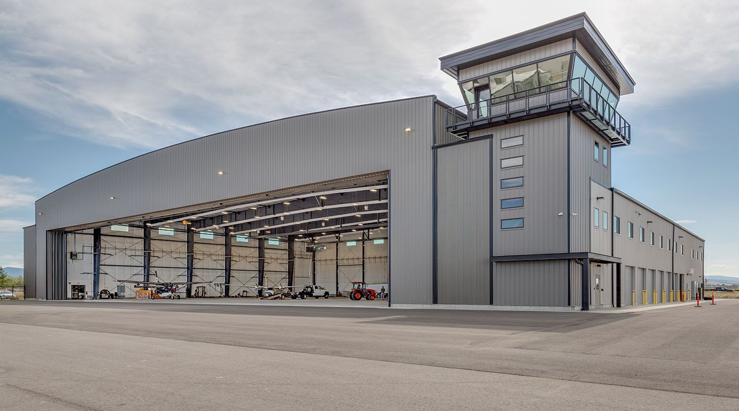 A large, modern gray industrial hangar building with a control tower on the right side under a cloudy sky