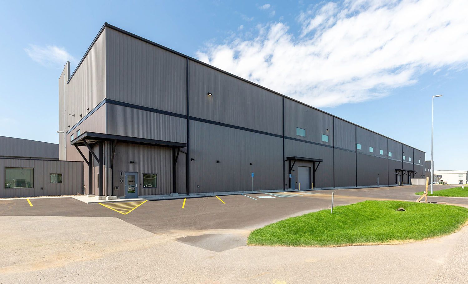 A large, modern, gray industrial warehouse building with a paved parking lot