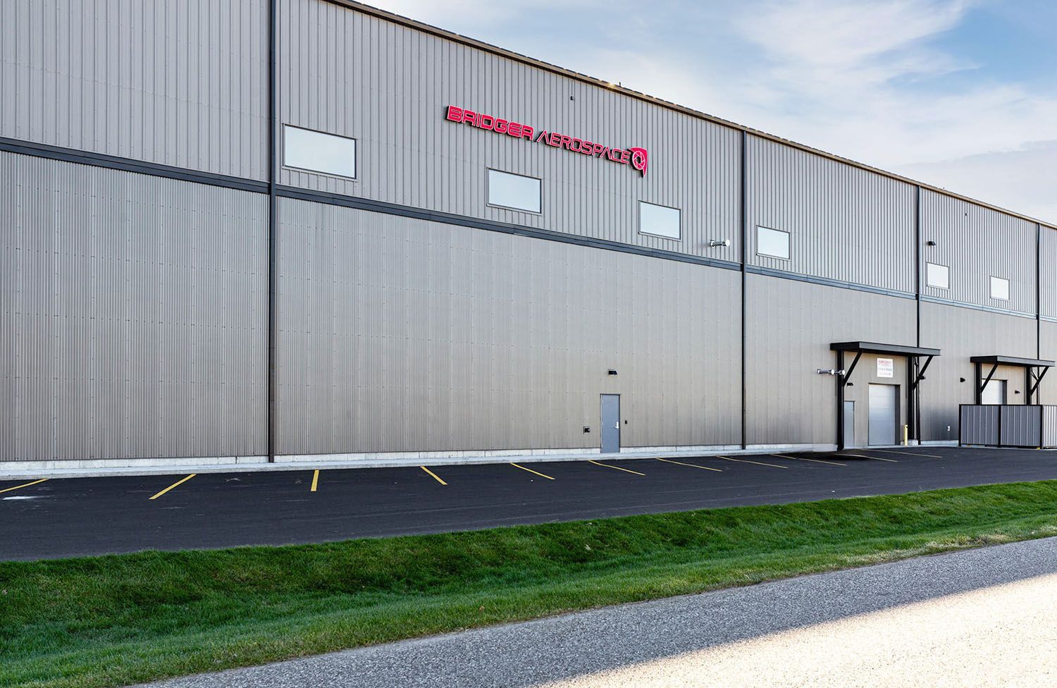 A grey, corrugated metal industrial building with a red sign, black parking spots, and a green grass strip in front