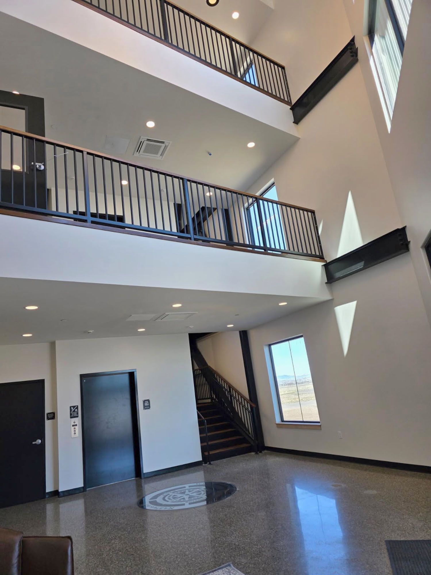 A modern two-story lobby featuring white walls, a metal balcony railing, a dark door, an elevator, and a staircase