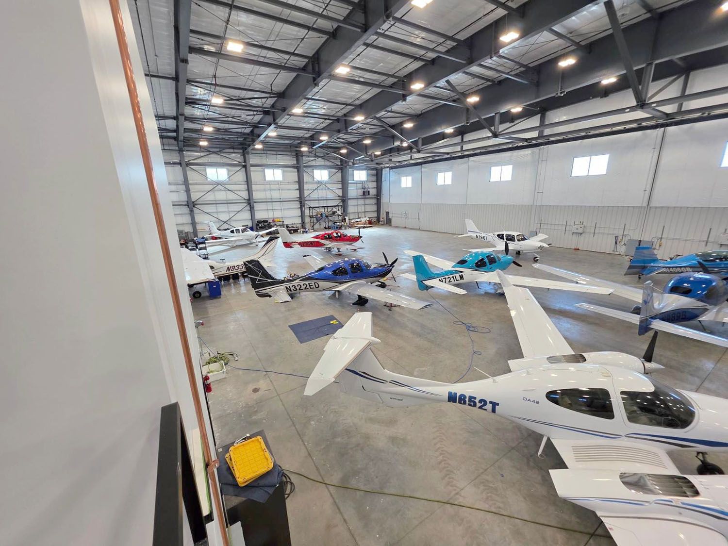 A large, brightly lit hangar filled with several small white, blue, and red airplanes parked on a concrete floor