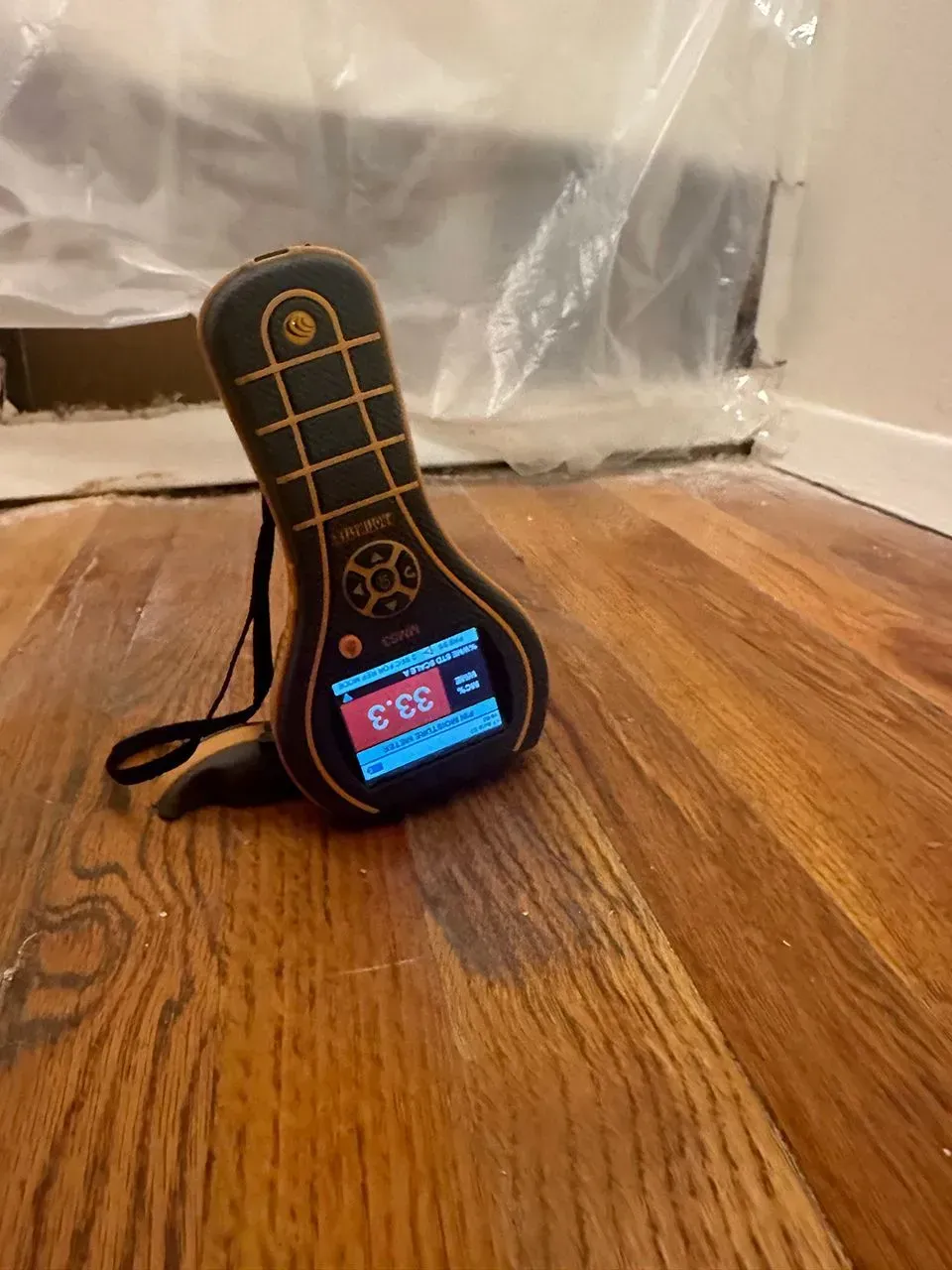 Moisture meter on hardwood floor, reading 33.3%, near wall with plastic sheeting. Orange and black device.