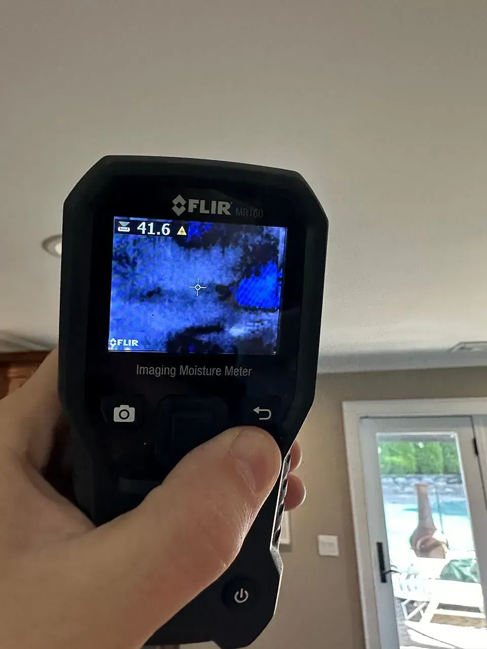Hand holding a FLIR thermal imaging camera pointed at a ceiling, displaying a blue area.