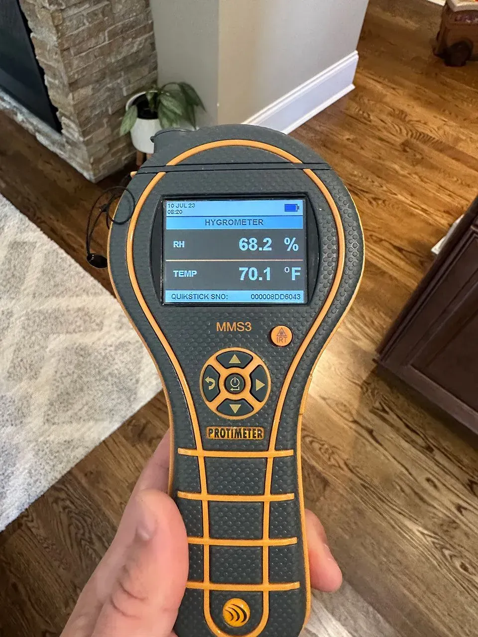 Hand holding a moisture meter displaying 68.2% humidity and 70.1°F temperature, in a room with hardwood flooring.