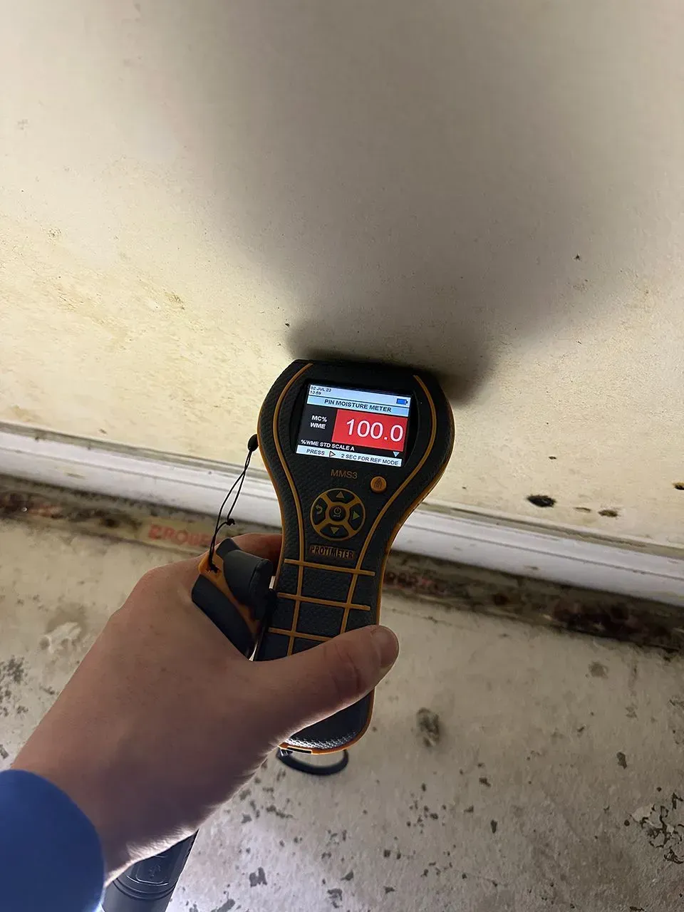 Hand holding a moisture meter measuring a ceiling, reading 100.0, in a room with stained walls.
