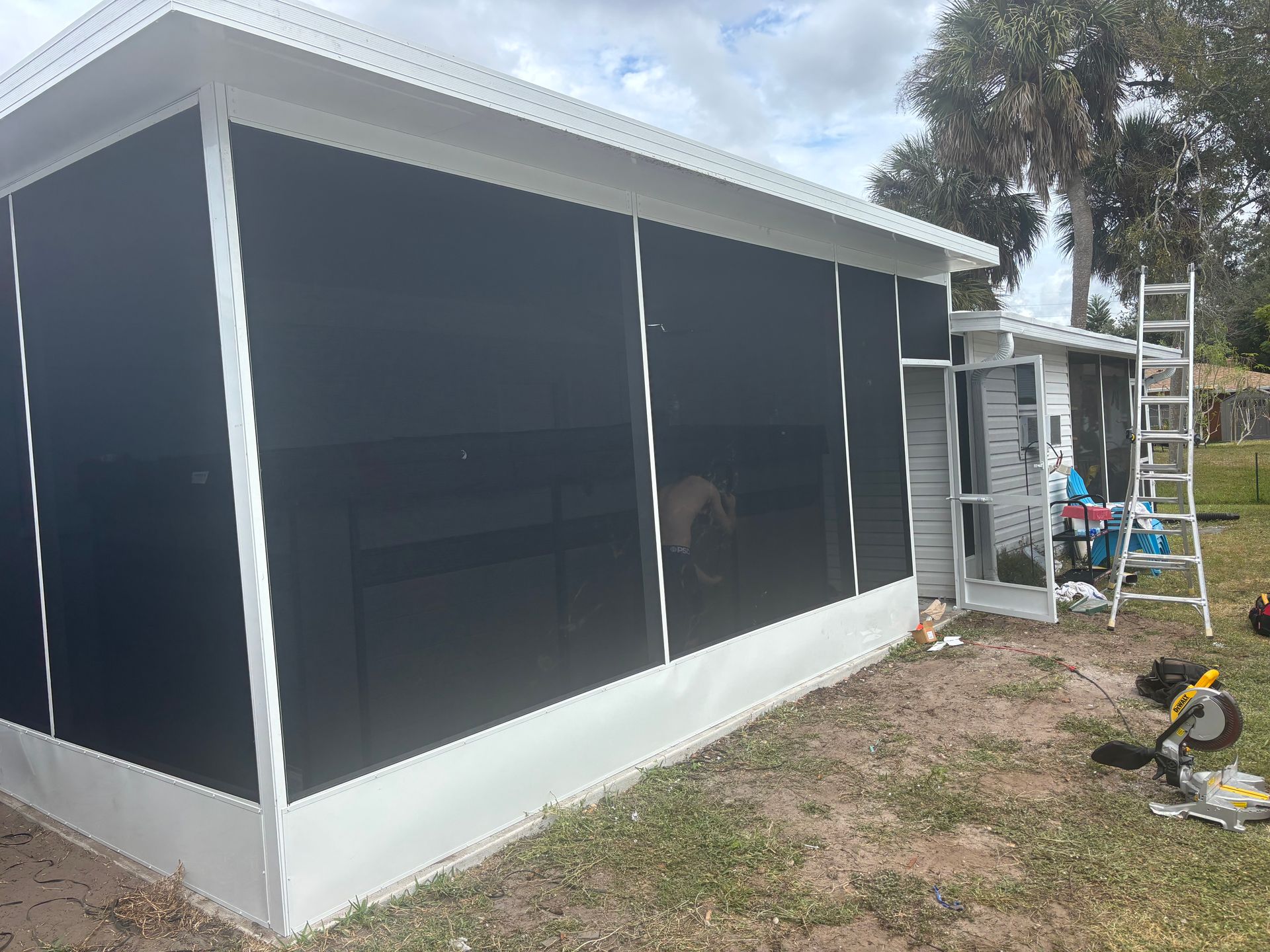 A white screen-enclosed patio with dark screens and a ladder, near a building on a grassy lawn.