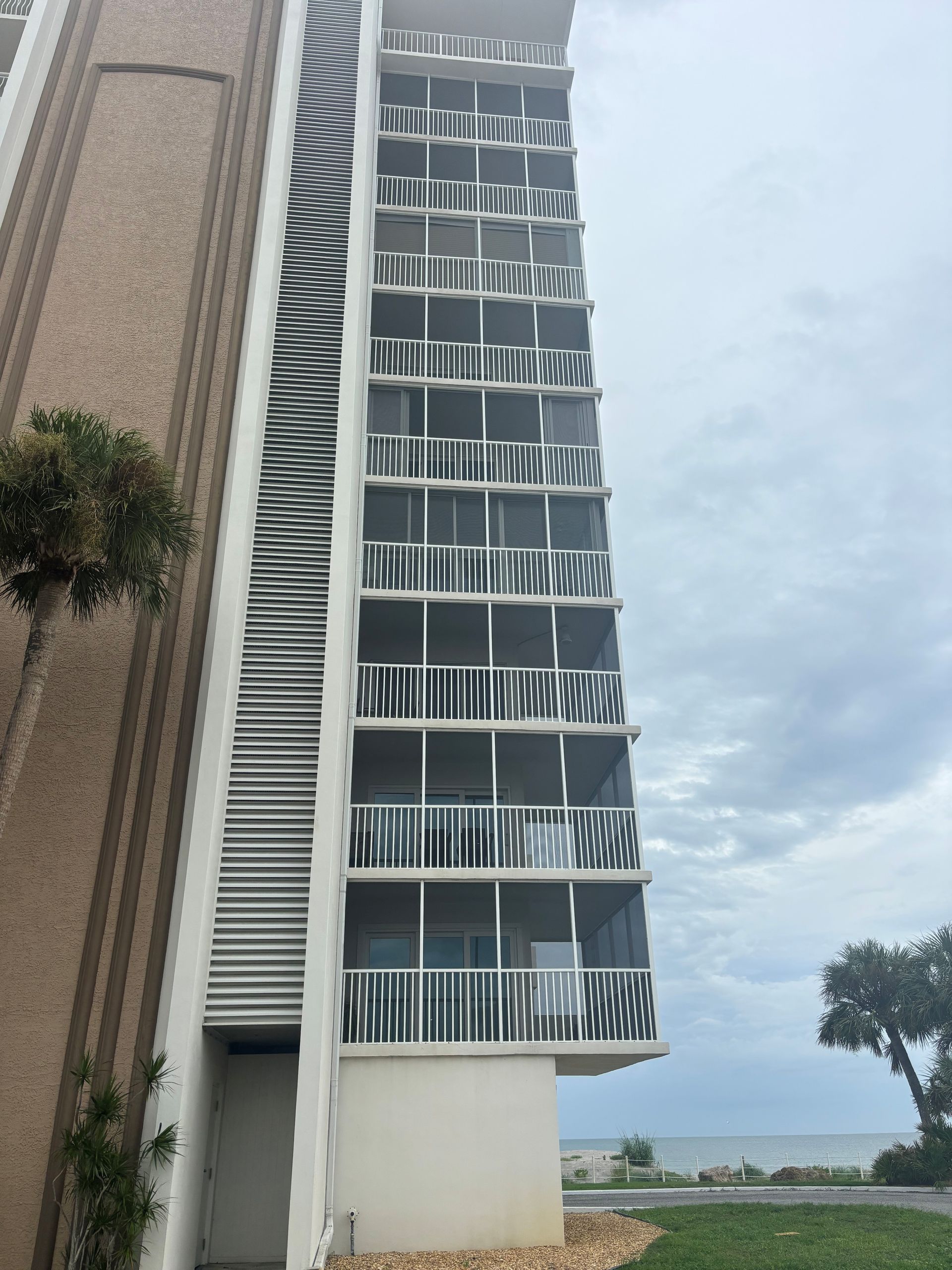 Tall building with stacked balconies facing the ocean. Overcast sky. Green grass in foreground.