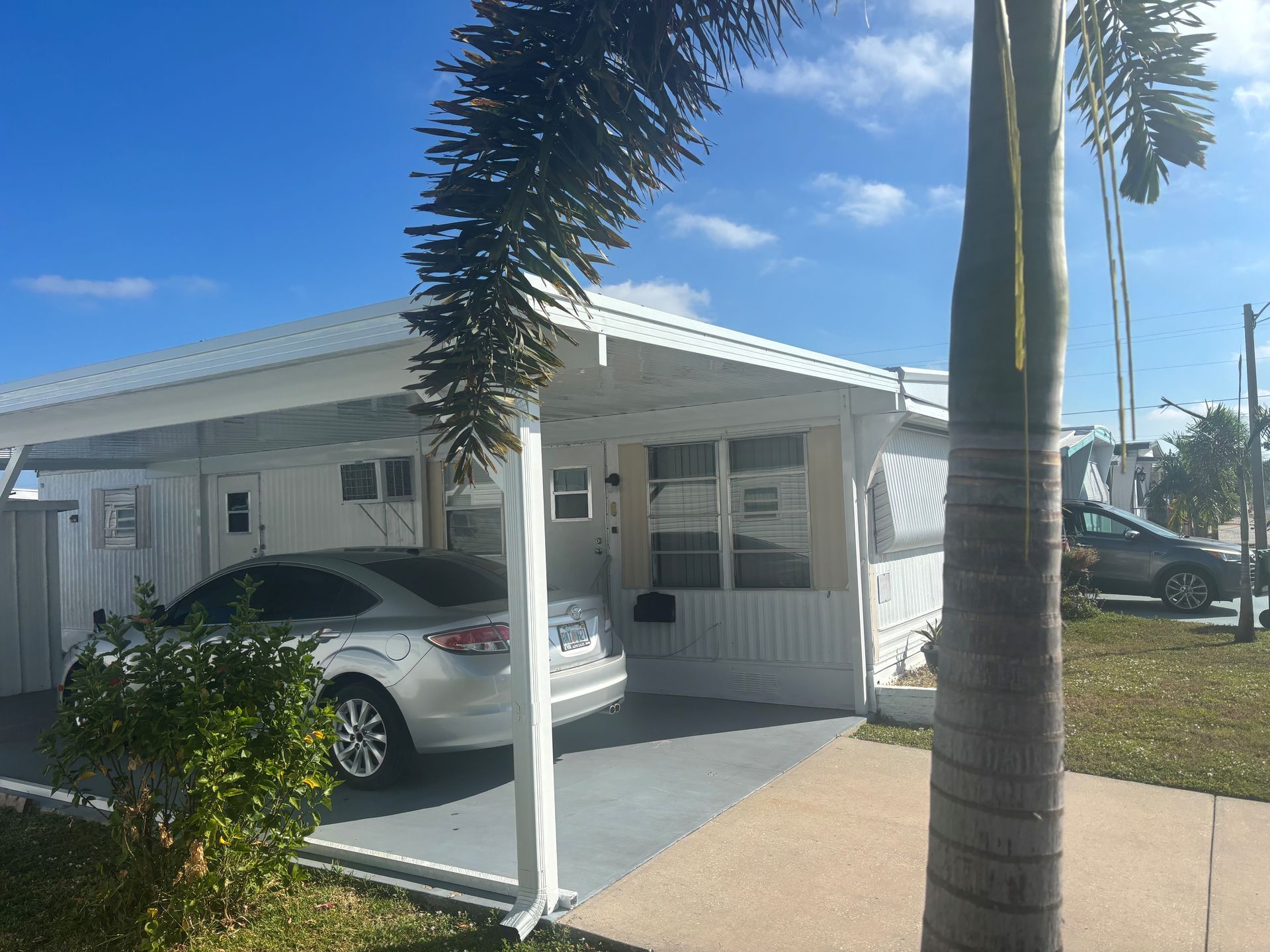 A silver car parked under a carport attached to a white mobile home. Palm trees and a blue sky are in the background.