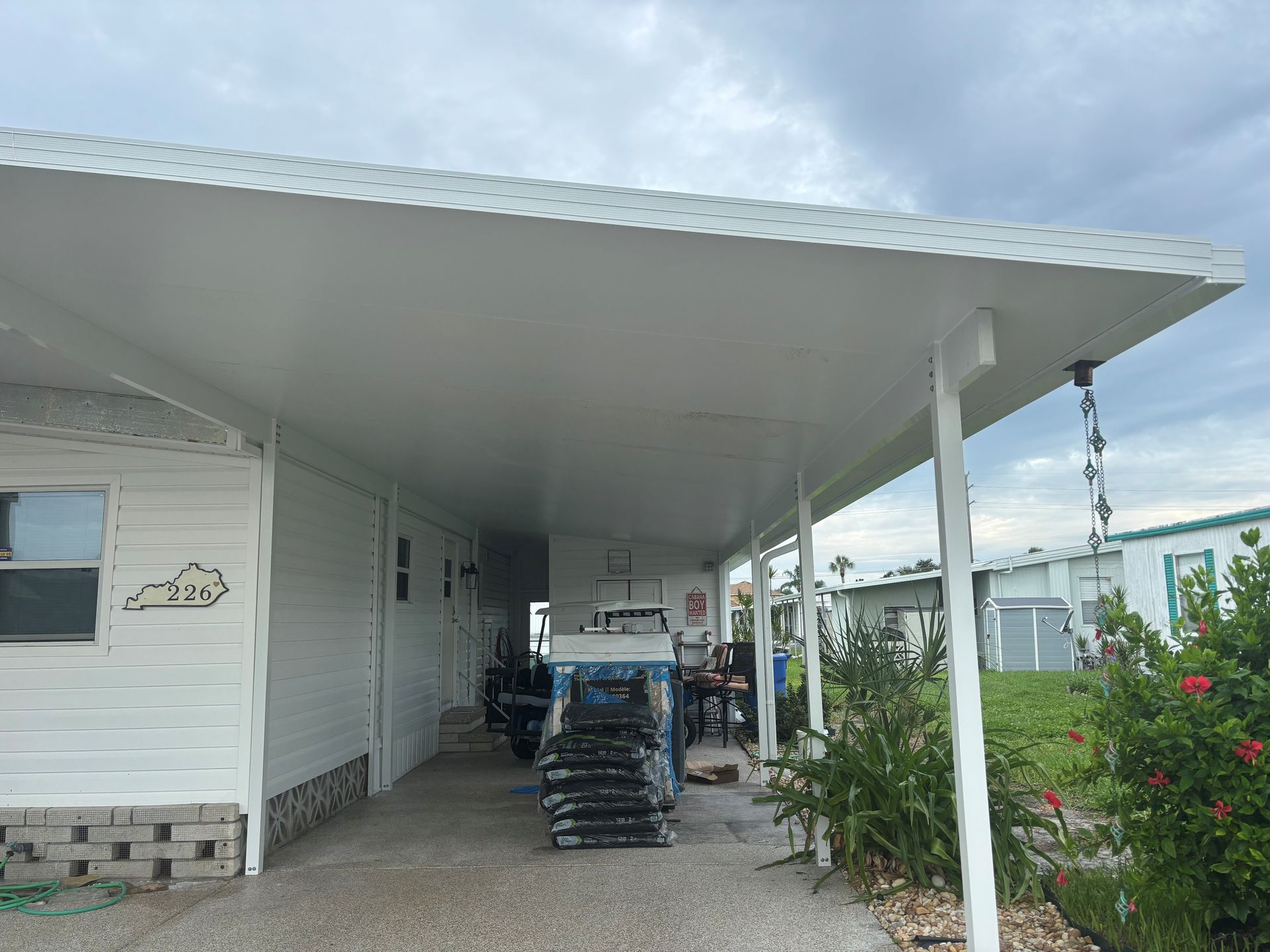 White carport attached to a mobile home, with items stored underneath. Overcast sky.