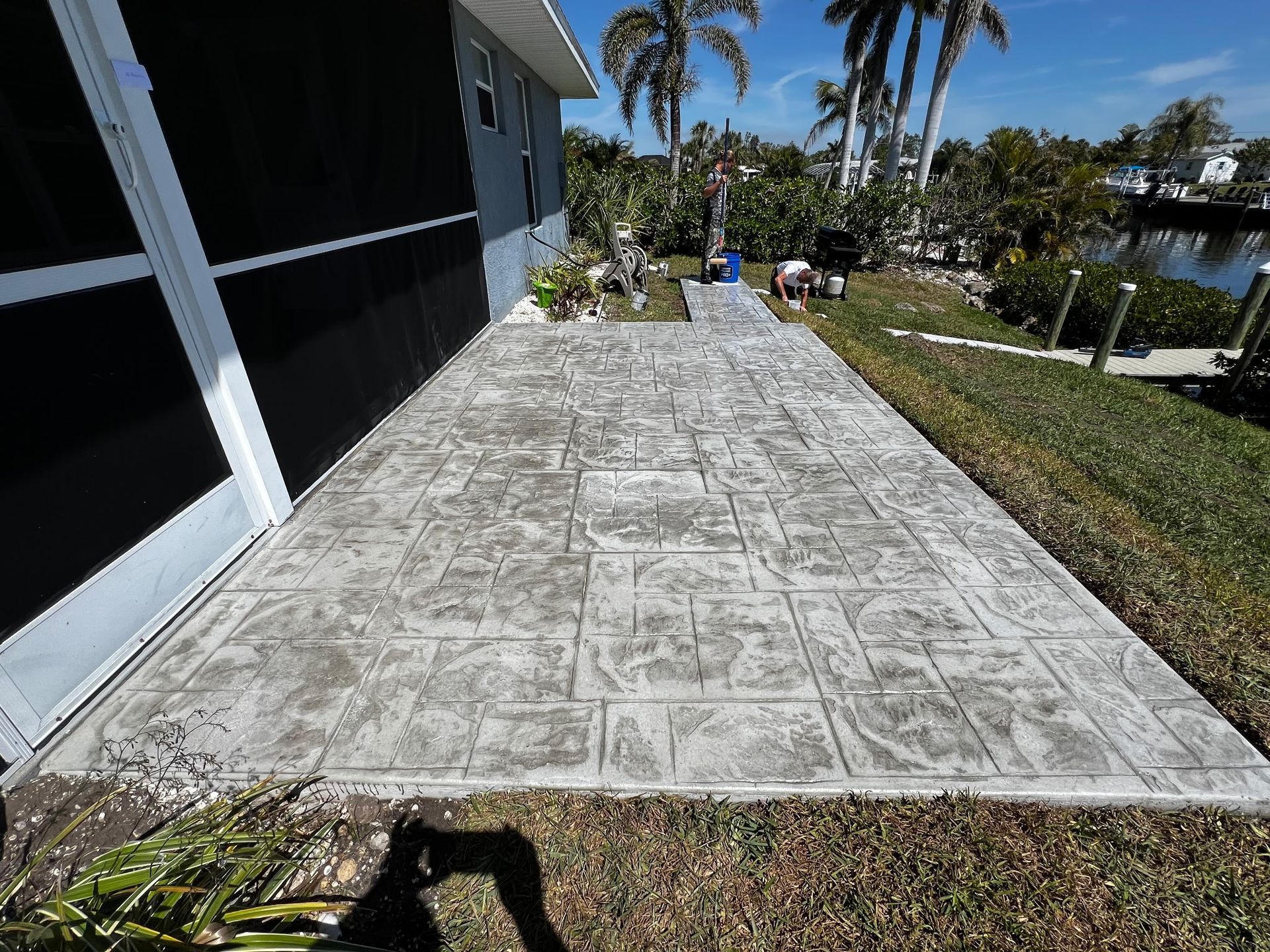 Stamped concrete patio adjacent to a light blue house, overlooking a canal with palm trees and a sunny sky.