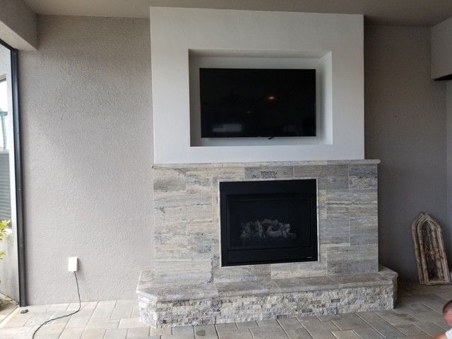Fireplace with a built-in TV above it. Stone facade, light colors, in an outdoor setting.