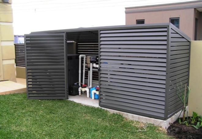 Dark gray slatted enclosure housing pool equipment, set on concrete, in a backyard.
