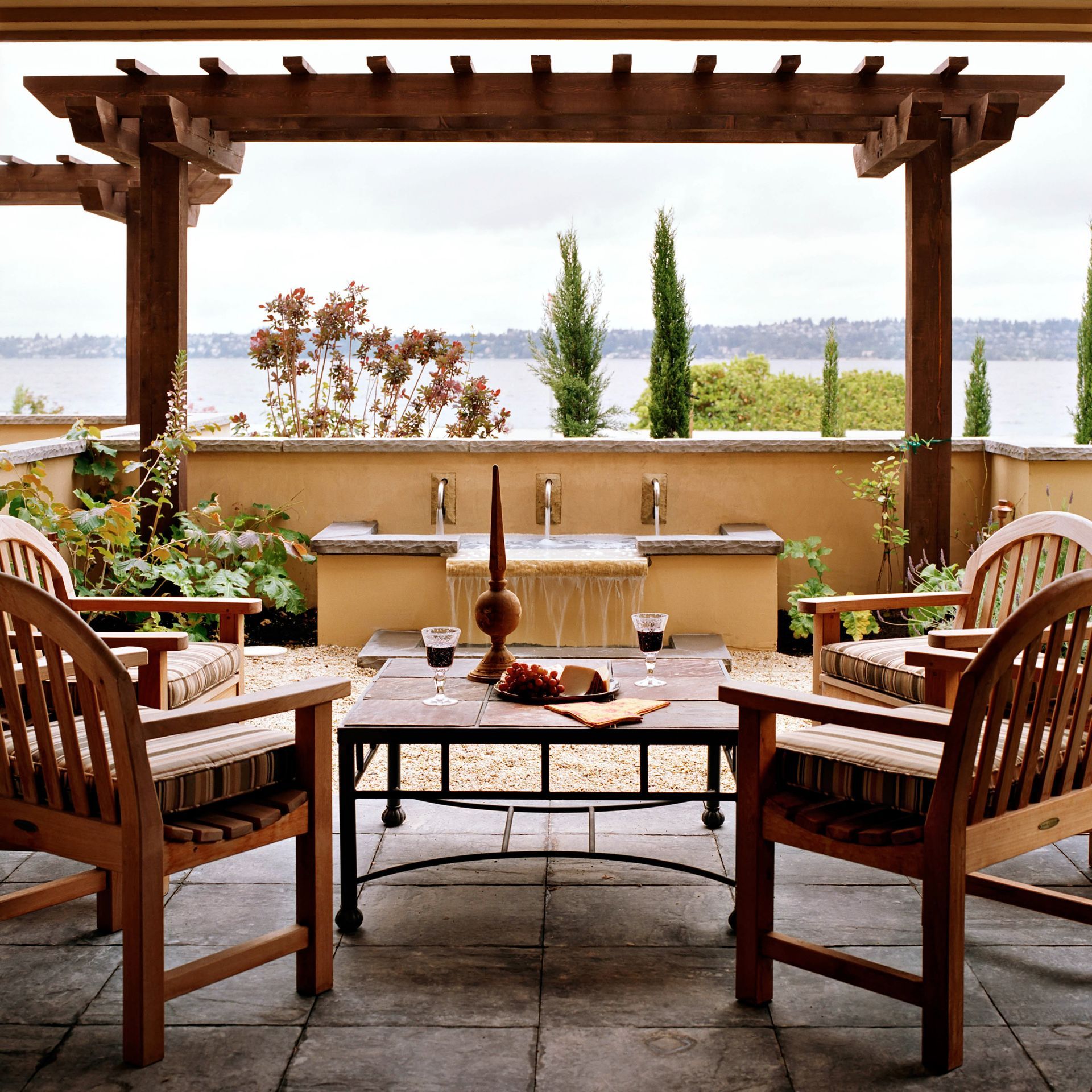 Outdoor patio with wooden furniture, pergola, fountain, and water view.
