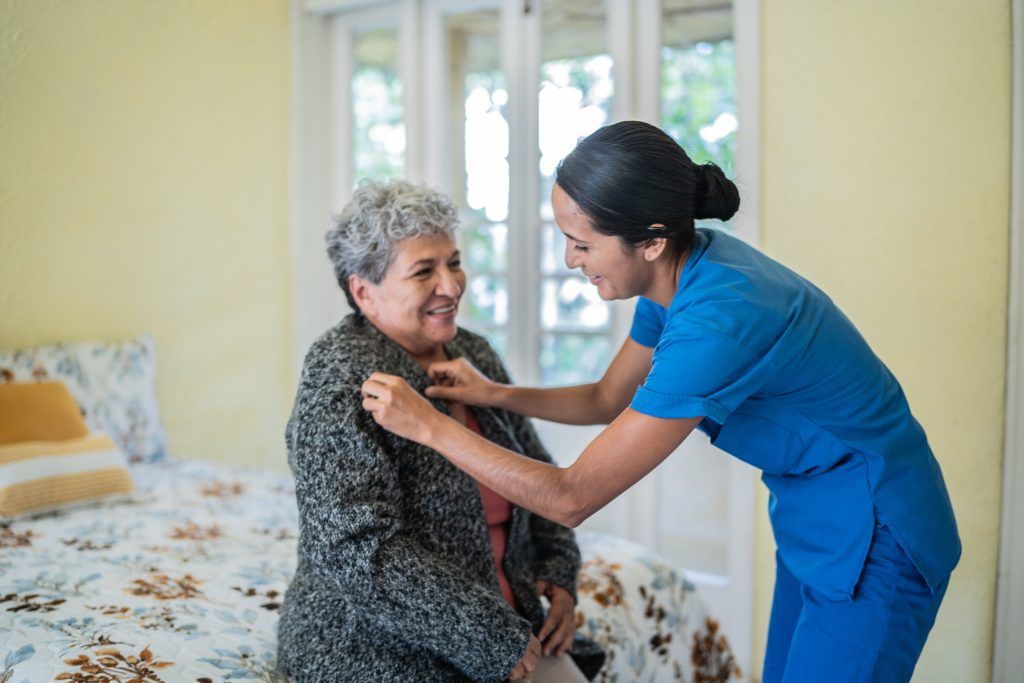 A nurse is helping an elderly woman dress in a bedroom.