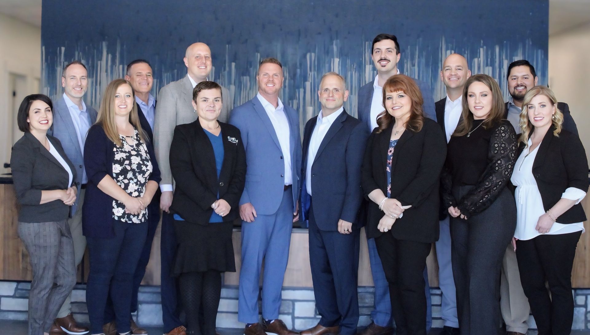Group of professionals standing together in front of a blue wall. They are diverse, mostly wearing business attire.