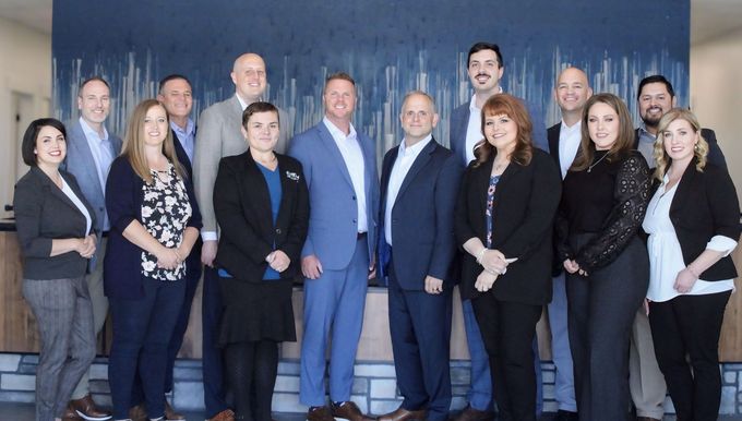 Group of professionals standing together in front of a blue wall. They are diverse, mostly wearing business attire.