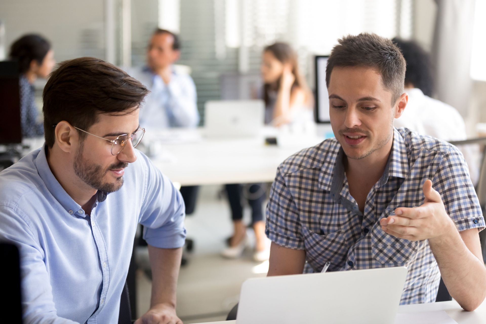 Two colleagues in a modern office discuss a project while looking at a laptop computer.
