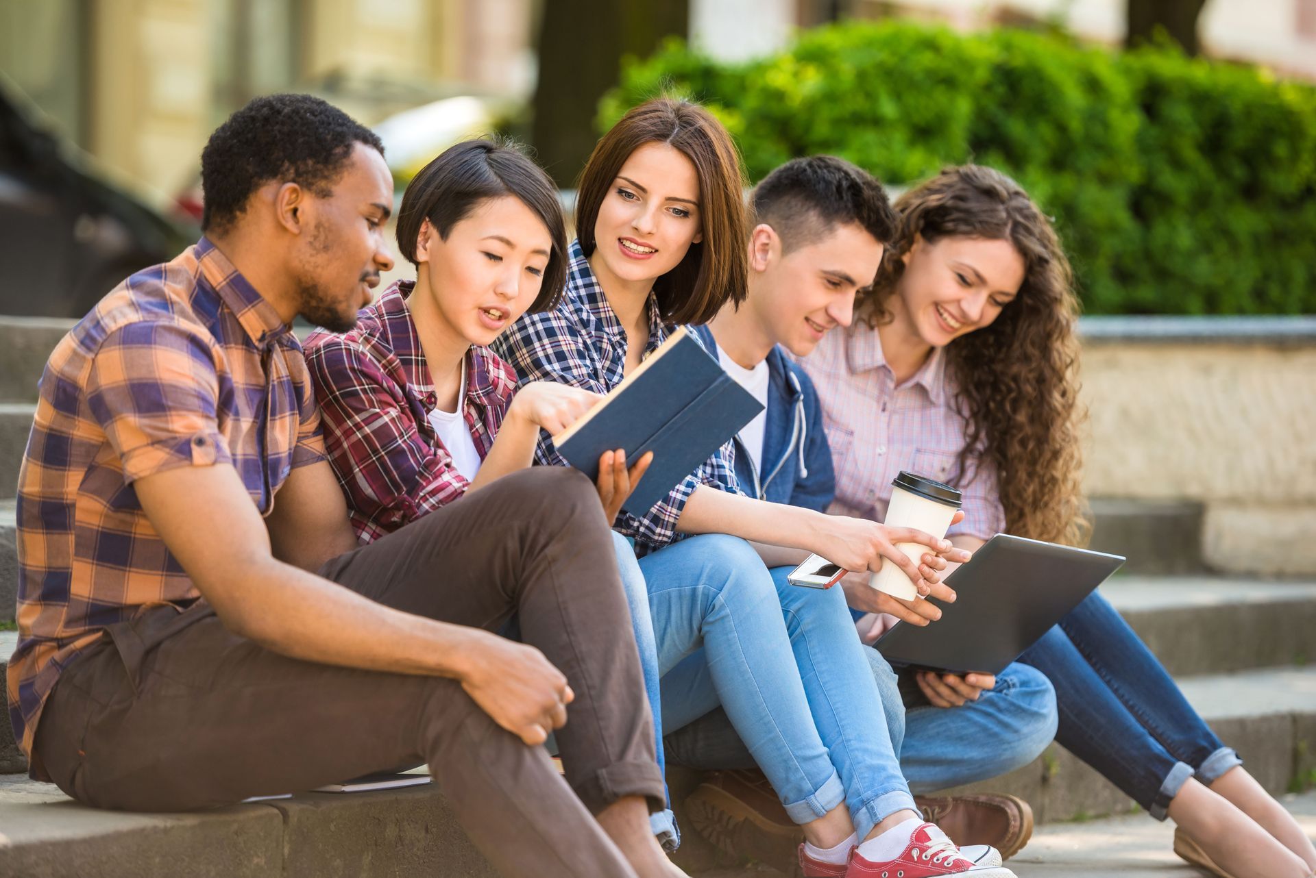 Five students sit together on outdoor steps, laughing and looking at a tablet and book while sharing a coffee.