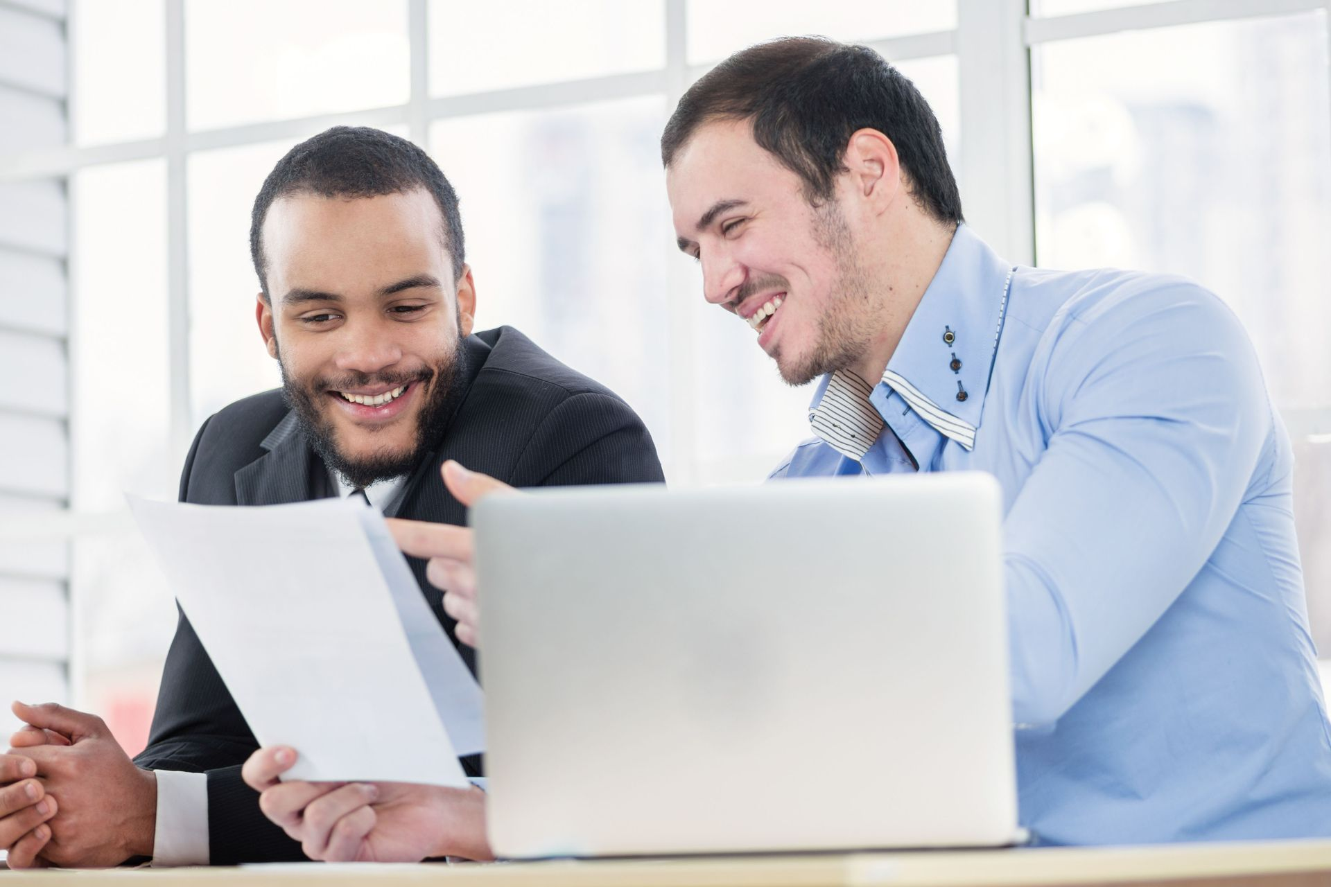 Two colleagues in a bright office look at a document together and smile, with a laptop open on the desk in front of them.