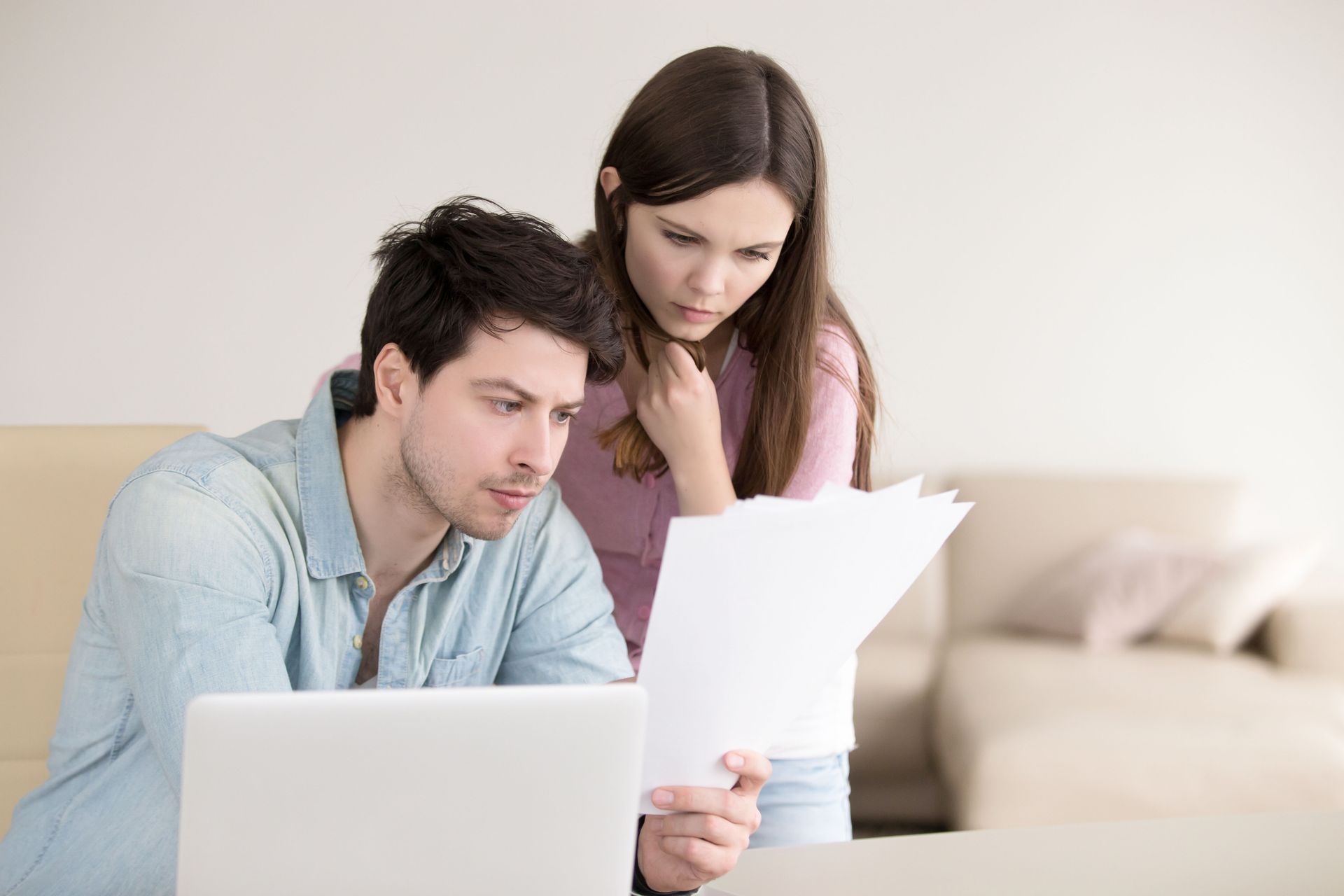 A person in a light blue shirt uses a laptop while another person holds a document, both looking at it with focus.