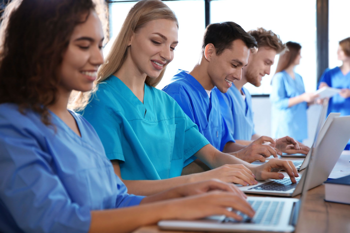 Medical students in blue scrubs working on laptops at a table, smiling.