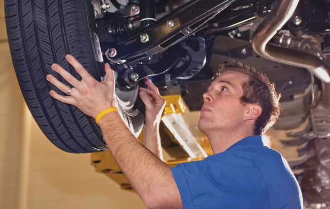 A mechanic in a blue shirt inspecting the underside and tire of a lifted vehicle in a garage.