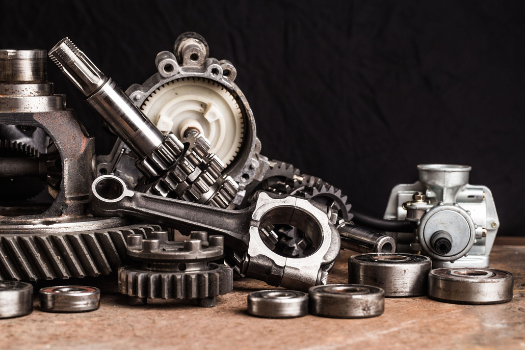 Assorted metallic engine parts, including gears, a connecting rod, and a carburetor, are scattered on a wooden surface.