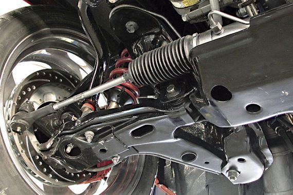 A low-angle view of a vehicle's front suspension, showing the control arm, steering rack, coil spring, and brake rotor.