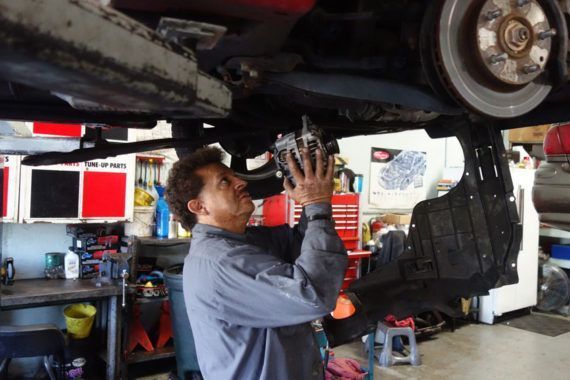 A mechanic in a grey shirt repairs a car engine while it is lifted on a hoist in an auto shop.