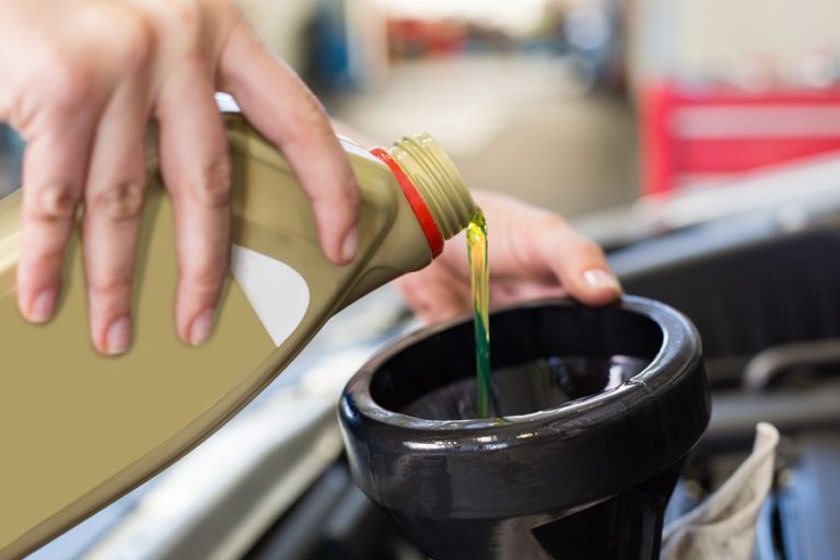 Hands pouring amber-colored oil from a plastic container into a black funnel in a car engine bay.