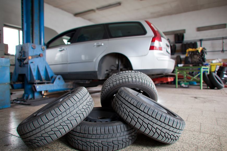 A pile of four car tires sitting on the floor in a garage with a silver station wagon lifted on a car lift in the back.