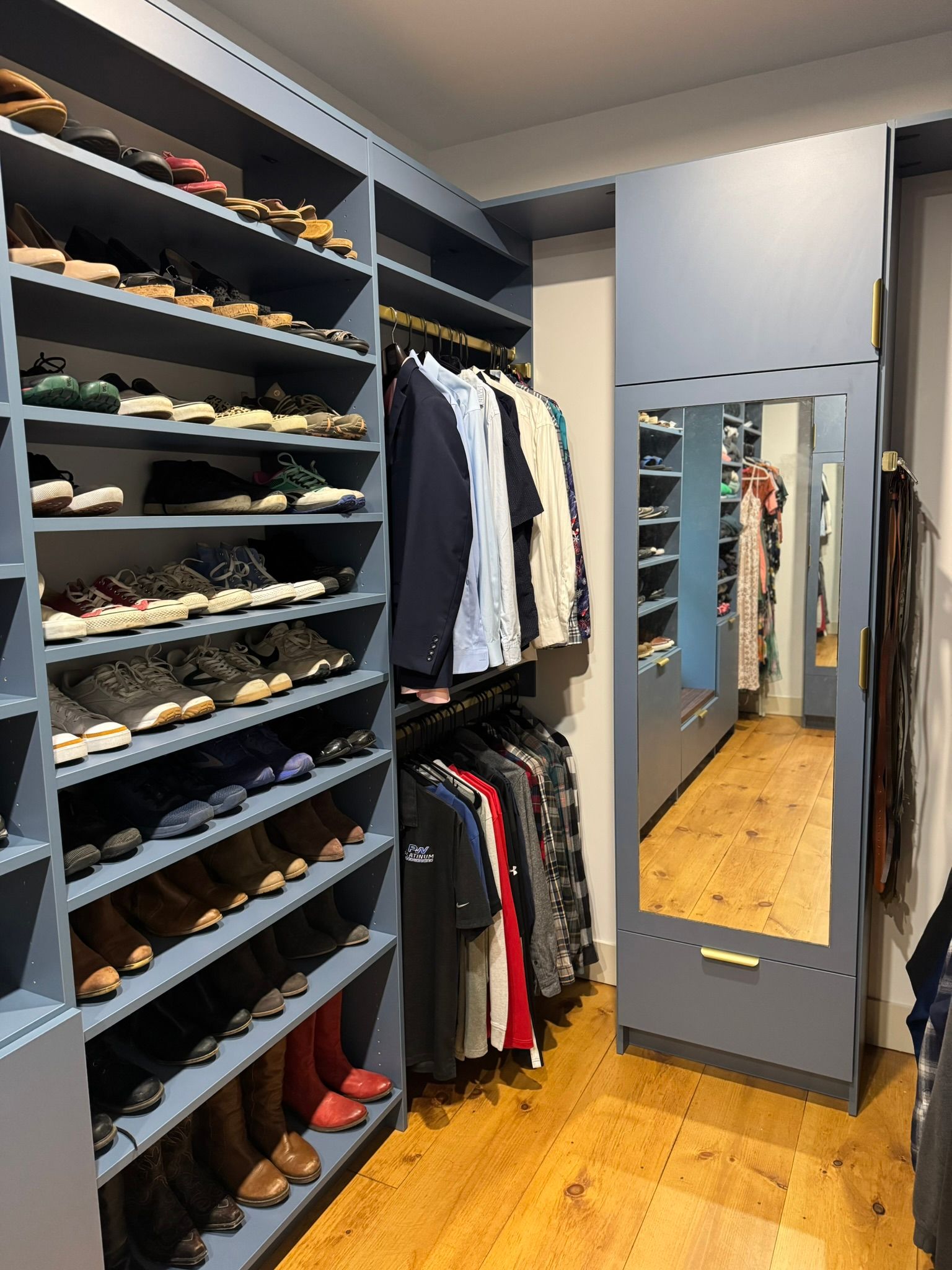 Walk-in closet with shoe shelves, hanging clothes, and a full-length mirror on a wooden floor.