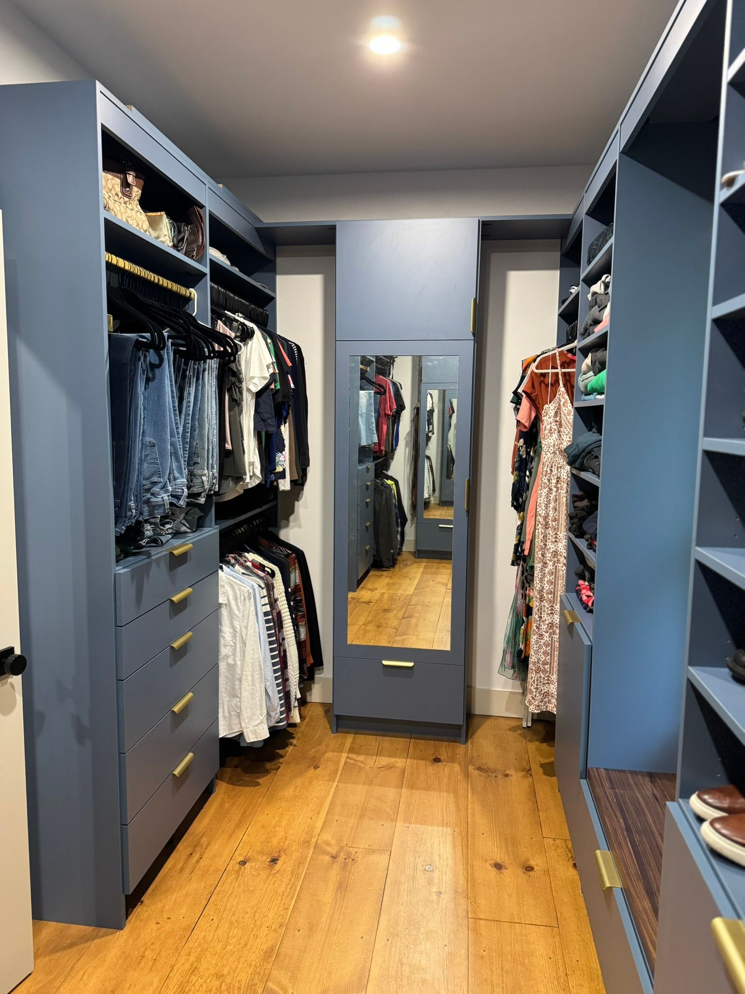 Narrow walk-in closet with blue shelving, hanging clothes, drawers, mirror, and wooden floor.