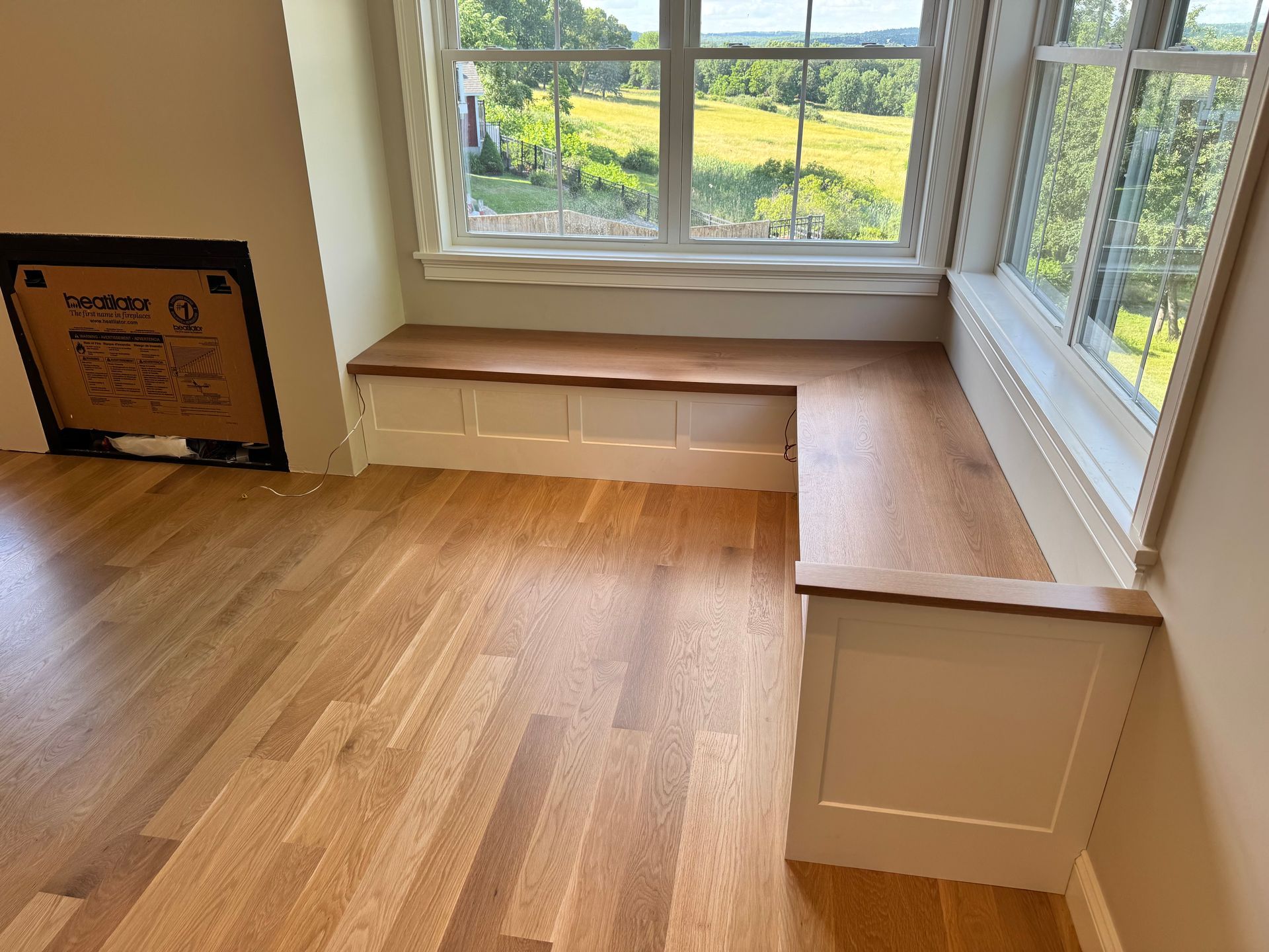 Bright room with wood floors and a window seat beneath a large window overlooking green fields.