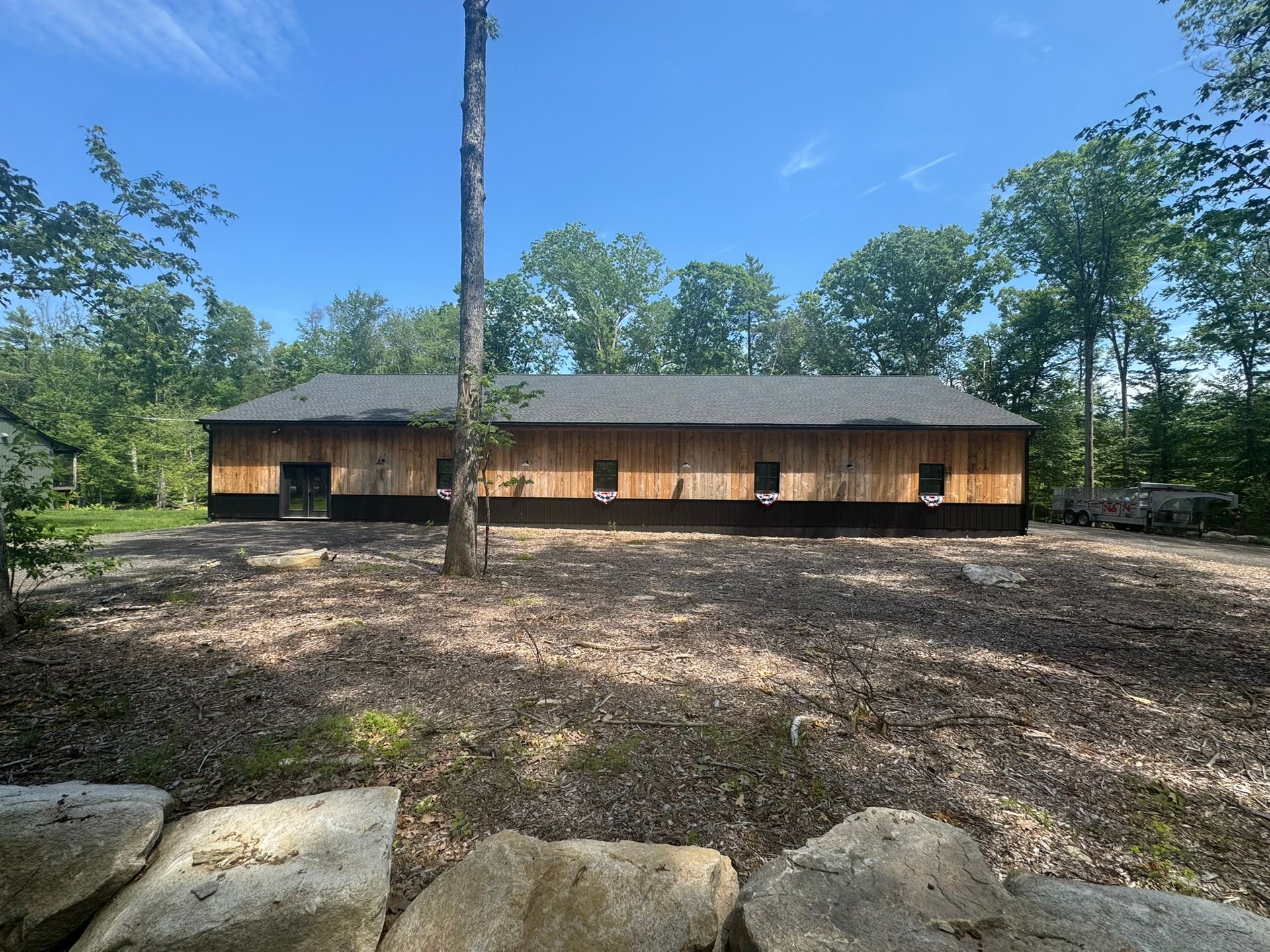 Long wooden cabin in a wooded clearing under a blue sky, with a large bare dirt foreground