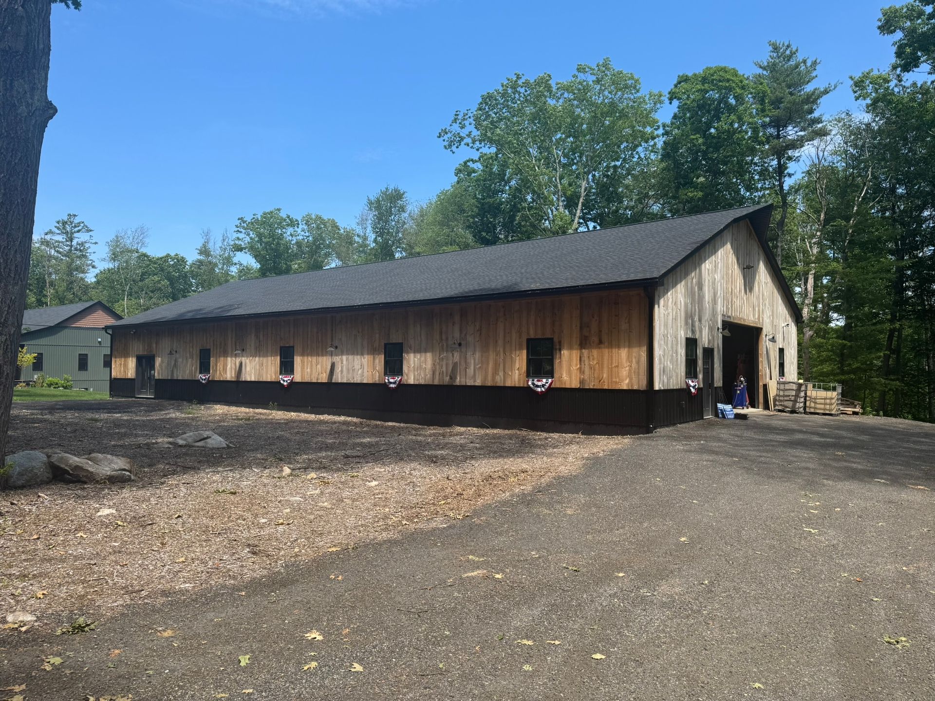 Long wooden barn with black roof on a gravel lot, surrounded by trees under a blue sky.