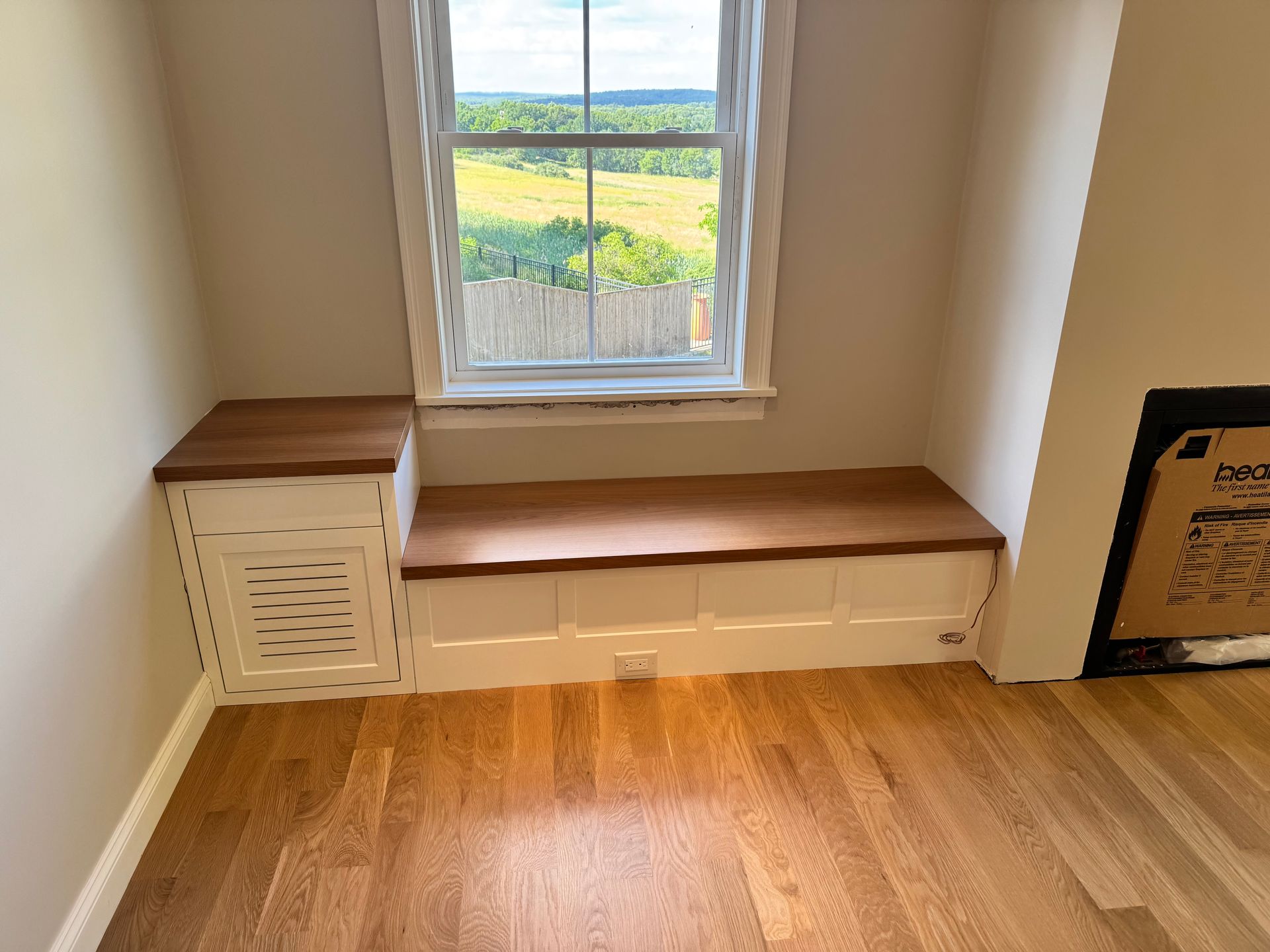 Built-in window seat with white storage cabinet in a bright room, overlooking green fields.