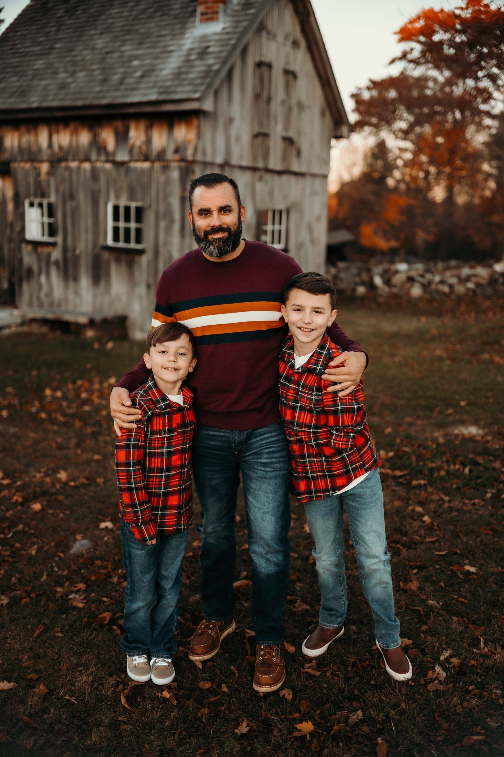 Man with two young boys in front of a wooden building; autumn colors, everyone smiles, and boy's wearing matching plaid shirts.