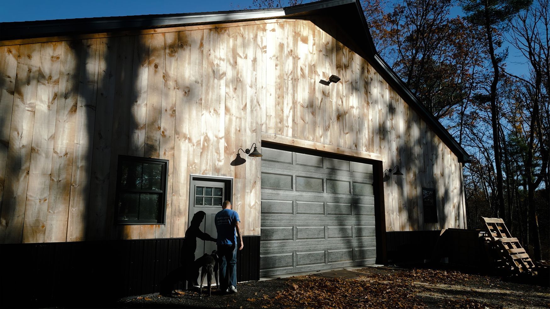 Barn with a large gray garage door, a small door, and two people standing outside. Light colored wood siding.