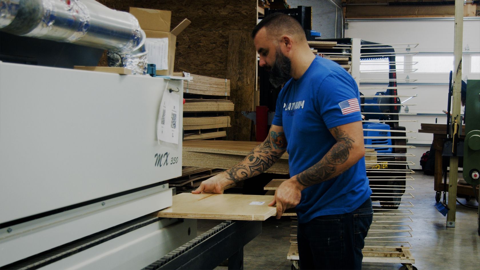 Man in blue shirt working with a large machine, holding a wooden panel in a workshop.