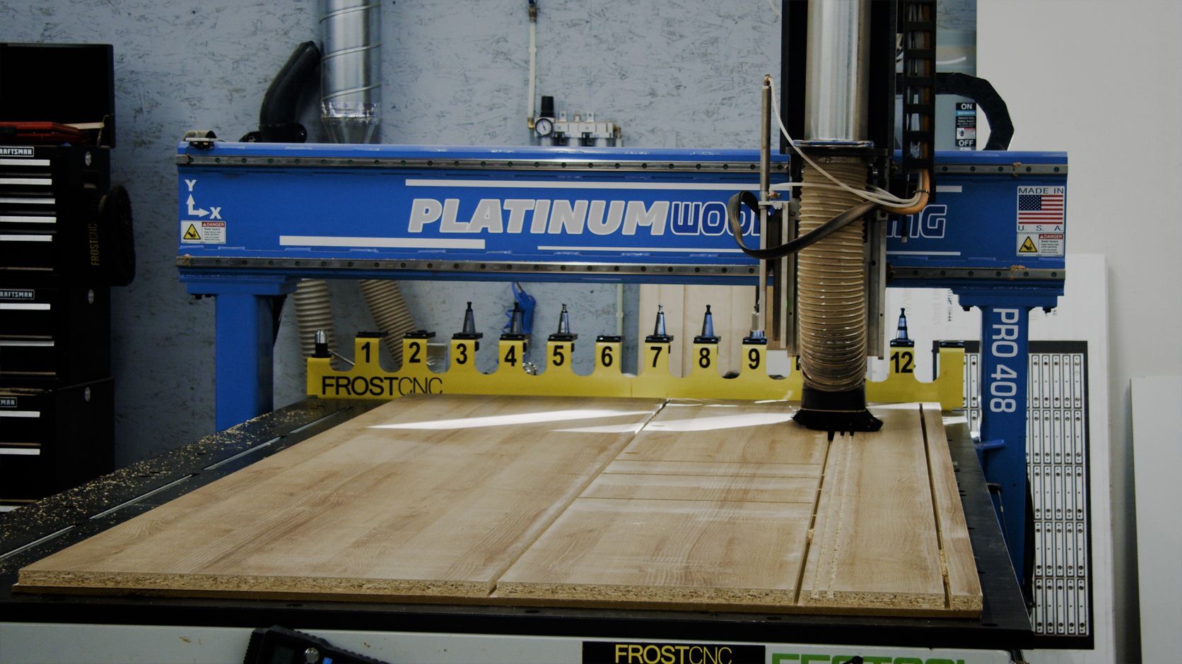 A CNC machine carving a pattern into a large wooden board. The machine is blue and yellow, in a workshop setting.