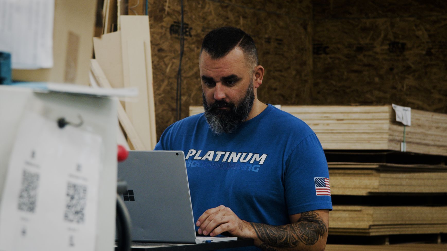 Man with beard typing on laptop in a woodworking shop; blue shirt, stacks of wood in background.