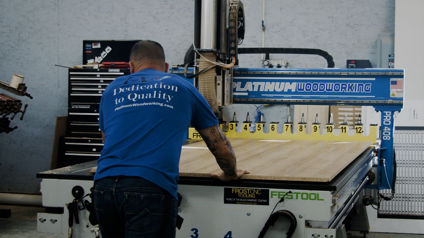 Man operates a CNC machine cutting wood in a workshop. Machine is blue, man wears a blue shirt.