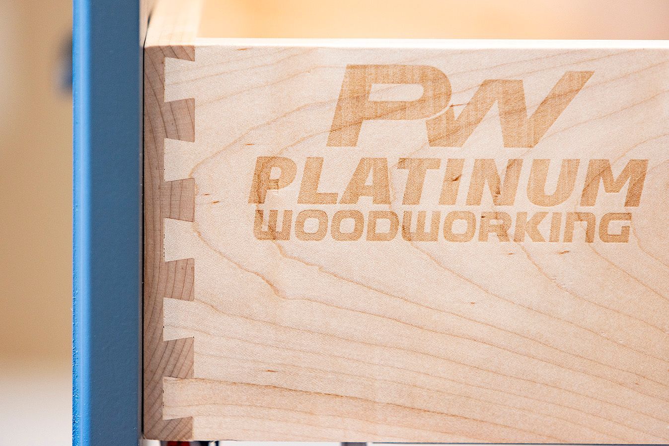 Close-up of a wooden drawer showing dovetail joints, the Platinum Woodworking logo, and a blue metal frame.