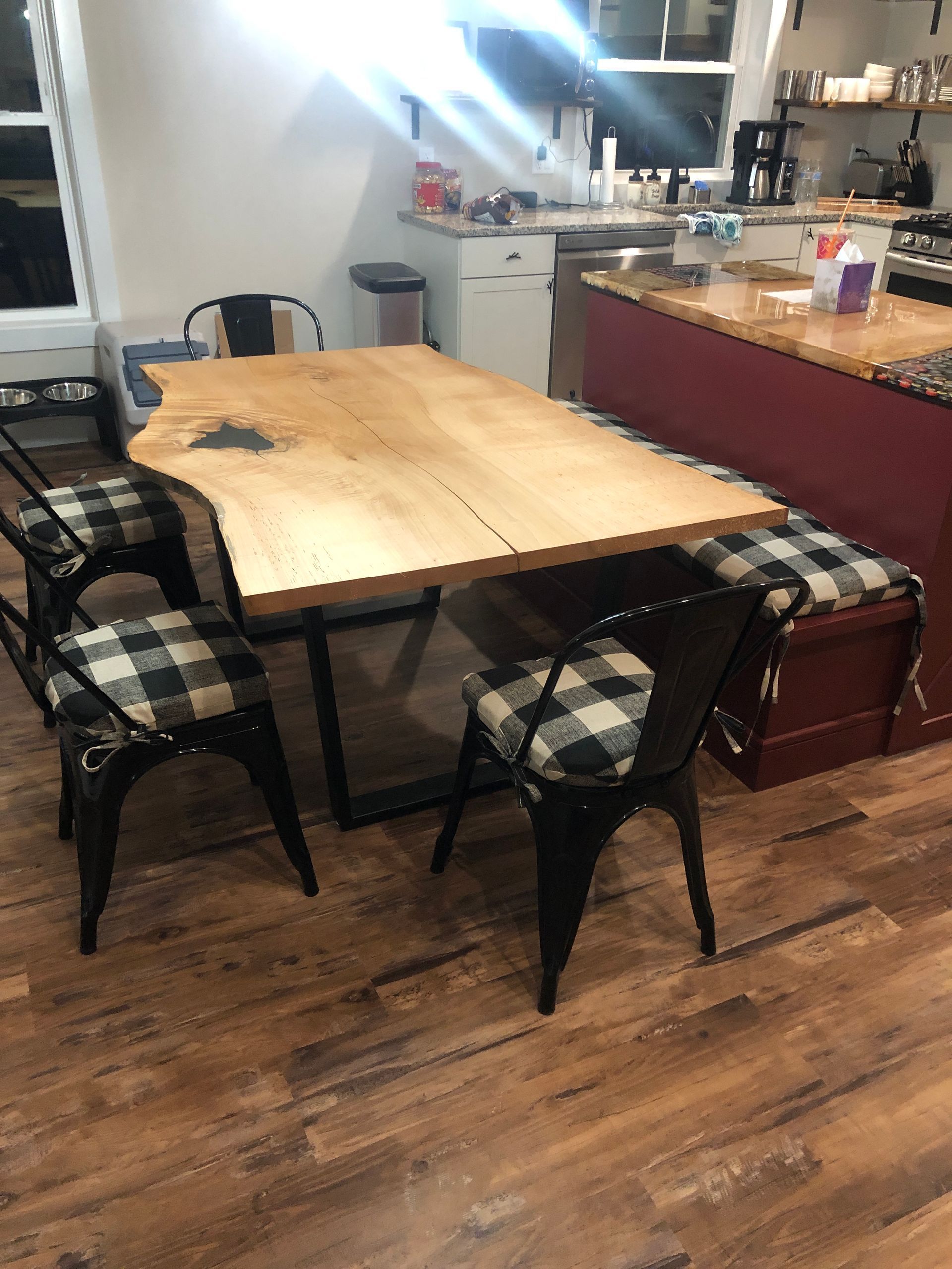 Wooden dining table with black metal chairs, plaid seat cushions, and a kitchen island in the background.